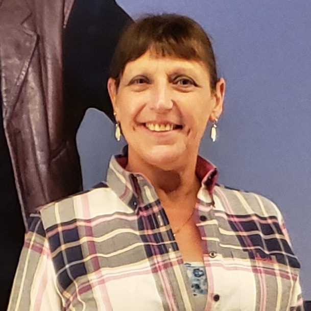 A person wearing a plaid shirt smiles at the camera, standing indoors against a blue and brown background.